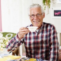 En vithårig och stilig Henry i röd-vit-blå-rutig skjorta sitter i sitt kök och dricker te
