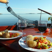 Två tallrikar med potatis och grönsaker uppdukade på bord på segelbåt bredvid två glas rött vin