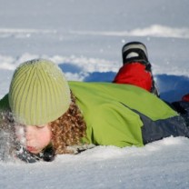 Lockhårigt barn med gröna vinterkläder och kängor ligger på mage i snön