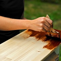 Kvinnohand håller en pensel mot en halvt brunmålad träplanka