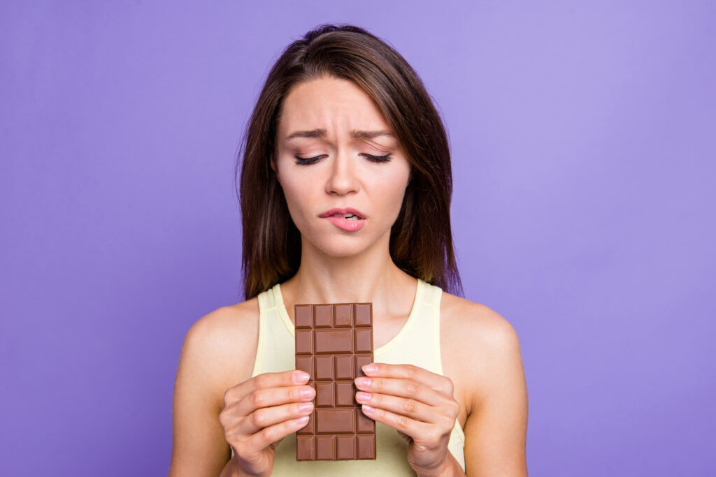 Close up portrait of lady with her brunette hair worried face she holds chocolate in hands look down bite low lip isolated on bright purple violet background