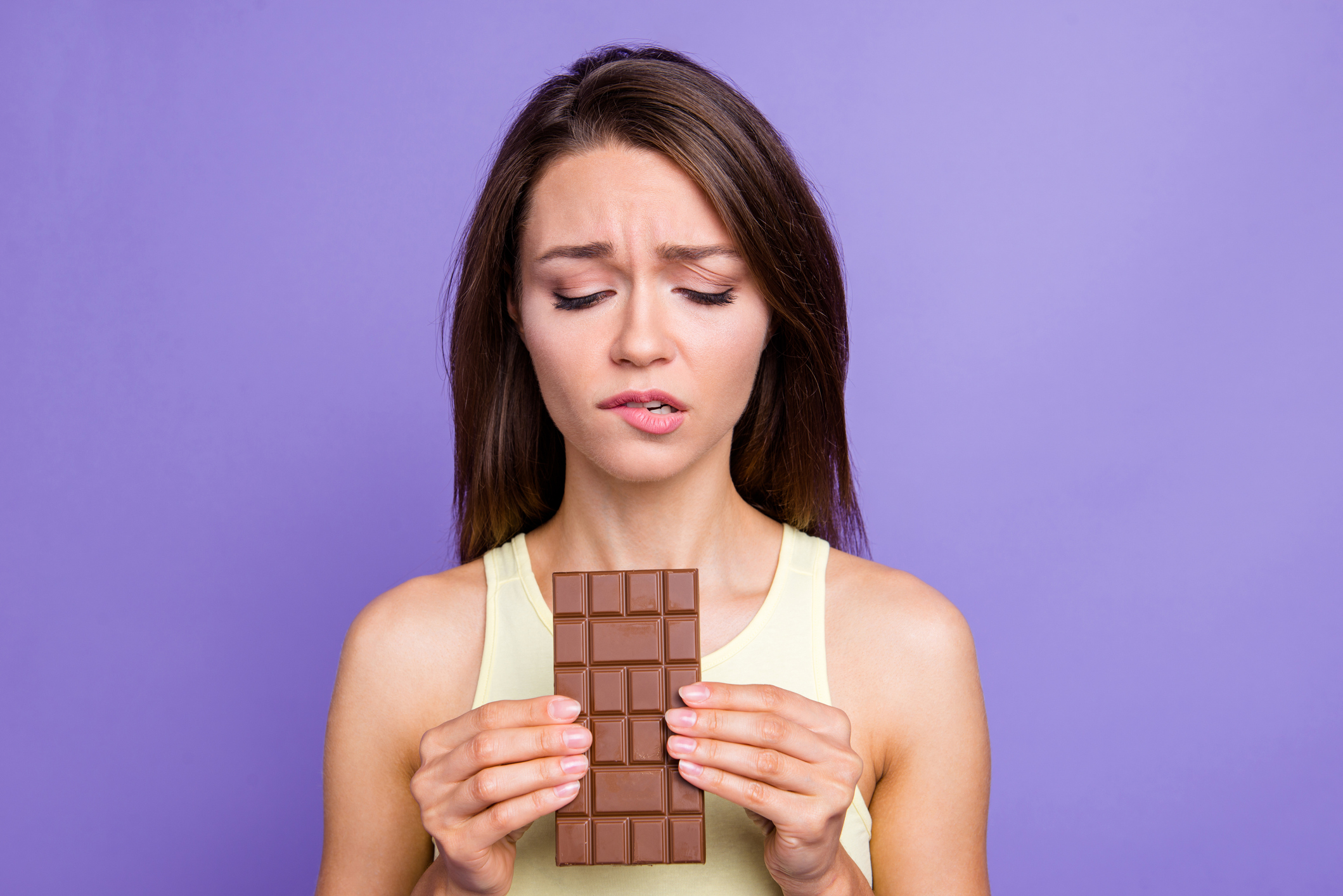 Close up portrait of lady with her brunette hair worried face she holds chocolate in hands look down bite low lip isolated on bright purple violet background