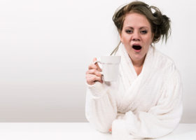 Trött kvinna i vit morgonrock gäspar med sin kaffe i handen