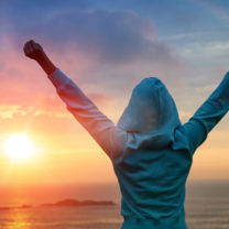 Kvinna håller upp händerna i luften i segerpose intill vatten och solnedgång
