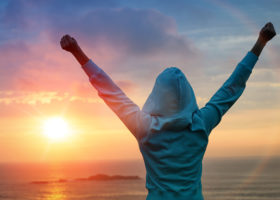 Kvinna håller upp händerna i luften i segerpose intill vatten och solnedgång