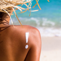 Solbrun kvinna vid vatten har ett utropstecken gjort av solskyddskräm på axeln