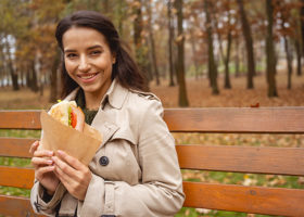 En kvinna i trenchcoat äter sin lunchmacka utomhus på en parkbänk