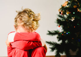 Pojke i tomtekläder sitter med ansiktet dolt i armarna framför en julgran