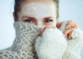 Kvinna med vit ansiktsmask som gömmer sig bakom en stickad polotröja