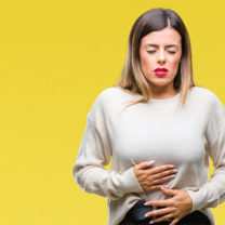 Kvinna med smärta och ont i magen, med handen på magen, gul bakgrund.