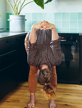 Kvinna står i yogaposition i köket med huvudet ner och händerna sammanflätade bakom ryggen