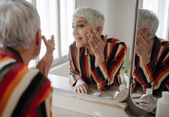 Kvinna med kort grått hår granskar sitt ansikte i spegeln