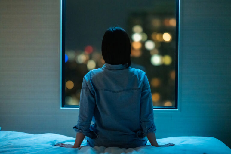 A rear view of a woman sitting alone on a bed in room and looking through the window at night.