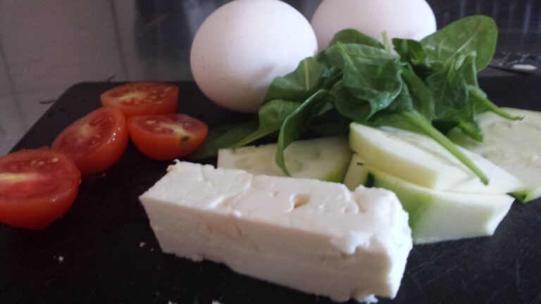 Ingredienser som tomat, fetaost, spenat och ägg till omelett på svart skärbräda