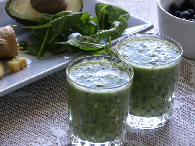 Två glas med grön smoothie. I bakgrunden ett grått fat med spenat, avokado och ingefära