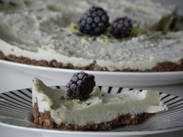 En bit Rawfood cheesecake på ett vitt fat med svarta ränder. I bakgrunden skymtar en hel cheesecake