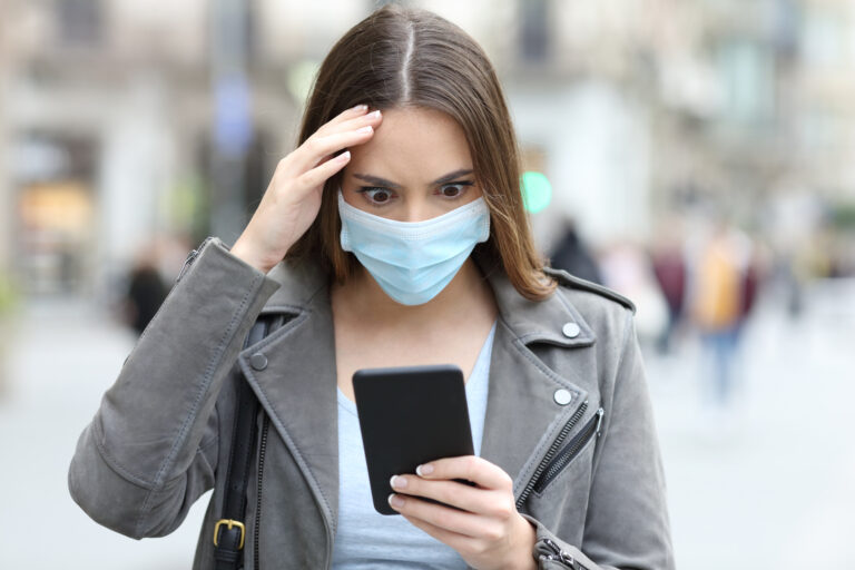 Ung kvinna med skyddande ansiktsmask ser orolig ut när hon ser på sin mobiltelefon