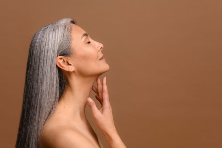 Profilbild mot brun medelålders kvinna med långt grått hår som tar på sin hud på halsen