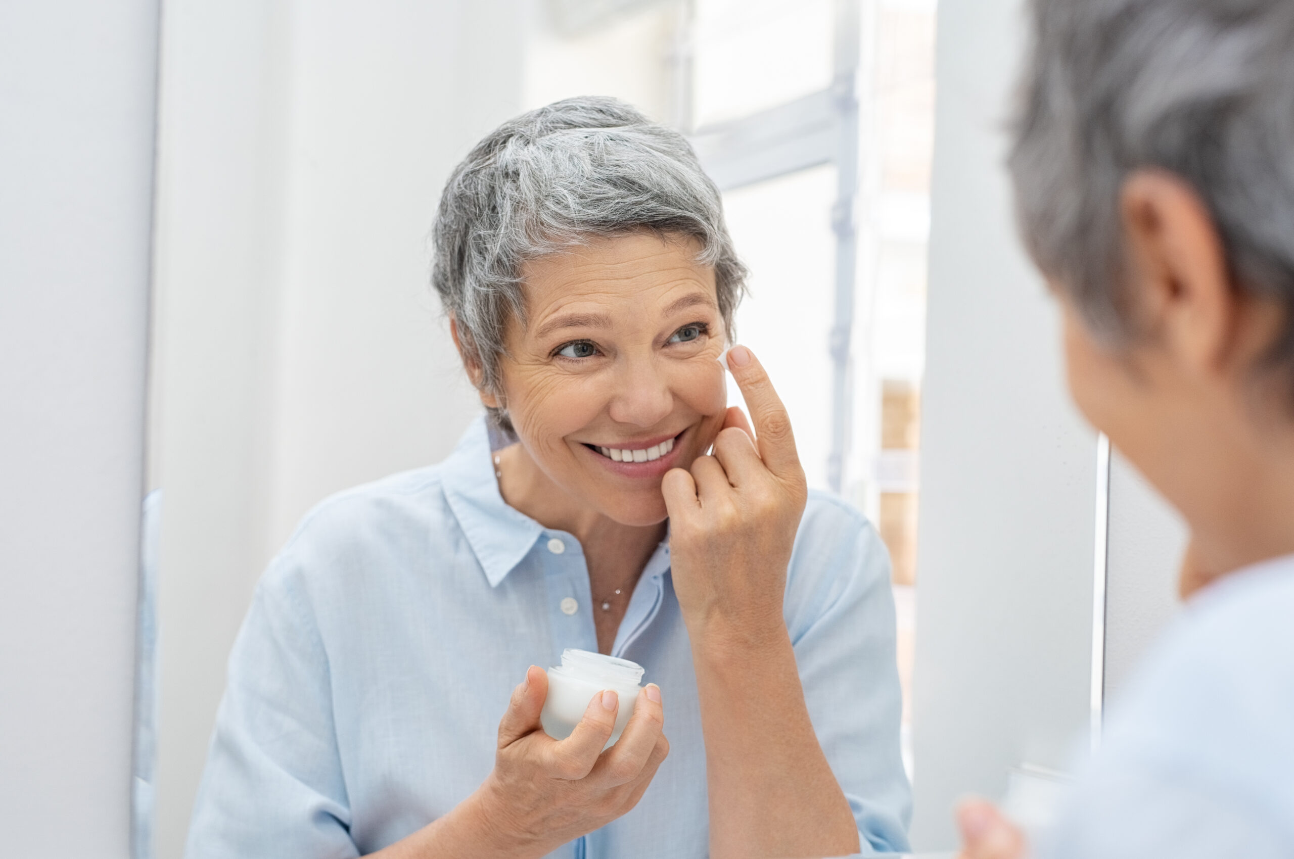 Happy mature woman applying face lotion while looking herself in the bathroom mirror. Senior woman applying anti aging moisturizer on her face. Smiling lady holding little jar of skin cream and applying lotion during the morning routine.