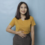 Pleased cheerful Asian woman keeps hand on belly feels full after delicious dinner dressed casually stands thoughtful against blue background.
