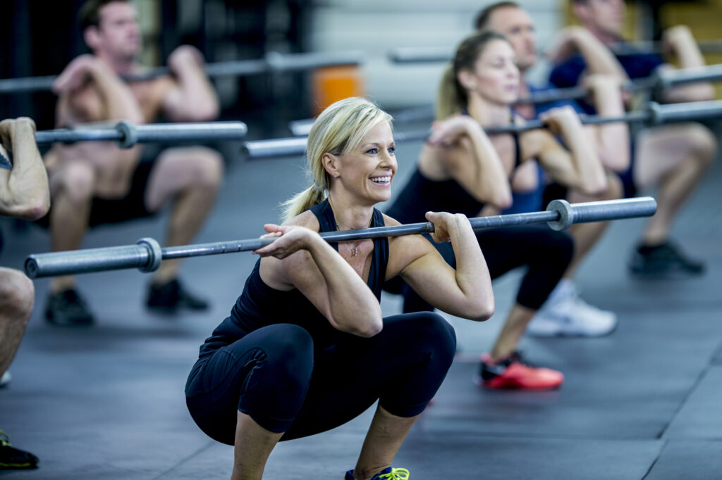 A group of adults are taking a fitness class together in a gym. They are wearing athletic clothes. They are squatting while holding a barbell.