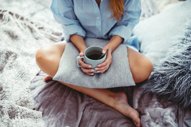 Närbild på kvinna i skräddarställning på säng med en kopp kaffe handen