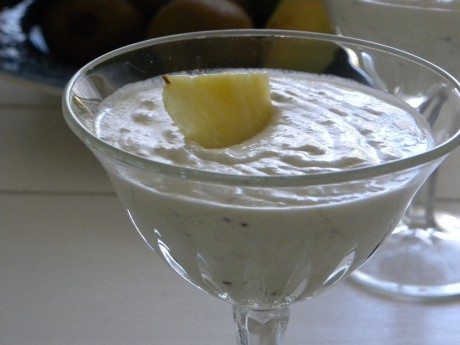 Kokossmoothie i glas toppad med ananas på vitt bord med frukt i bakgrunden