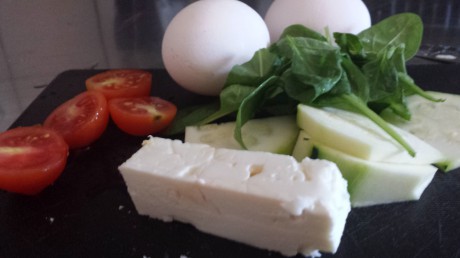 Ingredienser som tomat, fetaost, spenat och ägg till omelett på svart skärbräda