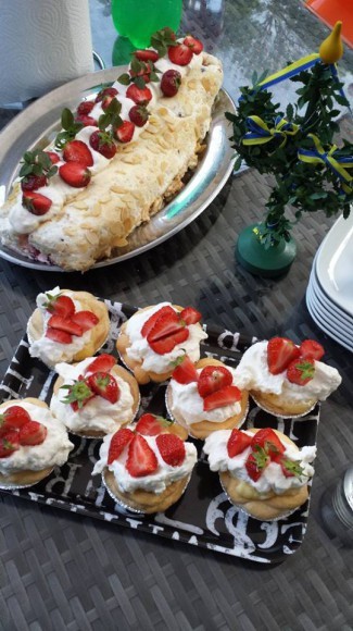 Budapestrulle med jordgubbar en midsommarstång i miniatyr och små bakelser med grädde och jordgubbar på ett bord